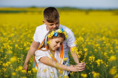 Mutlu kaygısız yerli çocukların ön görüntüsü: Genç erkek kardeş ve küçük kız kardeş, bulutlu gökyüzüne karşı sarı açan kolza tohumu tarlalarıyla çevrili yeşil çimlerle uzaklarda yürüyorlar.