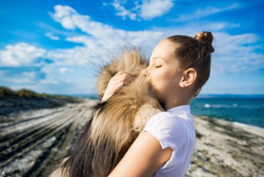 Saçları düzgün örülmüş sarı saçlı, beyaz tişörtlü, neşeli gülüşlü, pomeranian köpeğini öpen, altın sarısı uzun saçlı, Karadeniz kıyısında, güneşli rüzgarlı havada.
