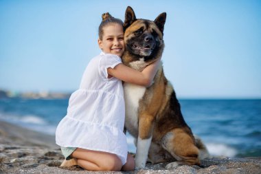 Hafif yaz kıyafetleri içinde neşeli mutlu genç kız sıcak ve güneşli havada Karadeniz 'in kıyısındaki geniş bir kumsalda Akita Inu soyunun tüylü, sadık, iri köpeğine sarılır ve onu sever.