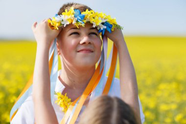 Nazik, genç kız kardeş, gülümseyen büyük erkek kardeşi, uzun renkli kurdeleli, parlak Ukrayna çelengini takıyor, sarı sınırsız tarlaların ve açık gökyüzünün arka planına karşı.