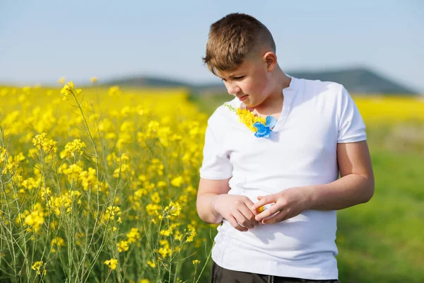 A big kind strong teenage guy in a white T-shirt with field Ukrainian wild flowers on it enjoys the warm summer bright sun while standing, in a yellow blooming rapeseed field against a blue clear sky