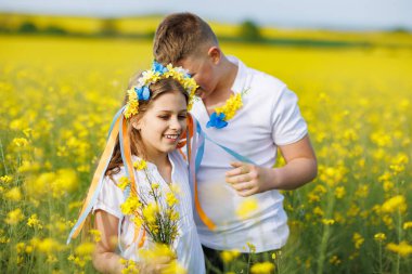 Mutlu kaygısız yerli çocukların ön görüntüsü: Genç erkek kardeş ve küçük kız kardeş, bulutlu gökyüzüne karşı sarı açan kolza tohumu tarlalarıyla çevrili yeşil çimlerle uzaklarda yürüyorlar.