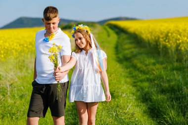 Mutlu kaygısız yerli çocukların ön görüntüsü: Genç erkek kardeş ve küçük kız kardeş, bulutlu gökyüzüne karşı sarı açan kolza tohumu tarlalarıyla çevrili yeşil çimlerle uzaklarda yürüyorlar.