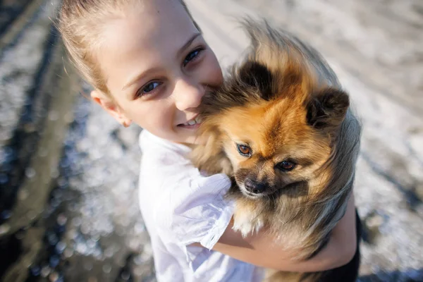 Saçları düzgün örülmüş sarı saçlı, beyaz tişörtlü, neşeli gülüşlü, pomeranian köpeğini öpen, altın sarısı uzun saçlı, Karadeniz kıyısında, güneşli rüzgarlı havada.
