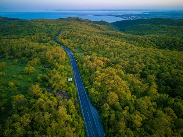 A winding new mountain road with fast modern cars and trucks leads into the distance, beyond the horizon, between green deciduous forests on the hills and the Balkan Mountains