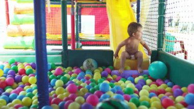 Küçük, kaygısız bir çocuk küçük bir oyun odasında oynuyor ve parlak küçük bir kayaktan renkli toplarla dolu bir havuza yuvarlanıyor. 4K UHD yavaş çekim videosu