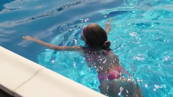 一个穿着鲜艳的粉色泳衣 戴着儿童紫色眼镜的美丽而滑稽的瘦小女孩 在一个温暖的阳光明媚的夏日 漂浮在一个巨大而又透明的游泳池里 清澈的水面上 — 图库视频影像