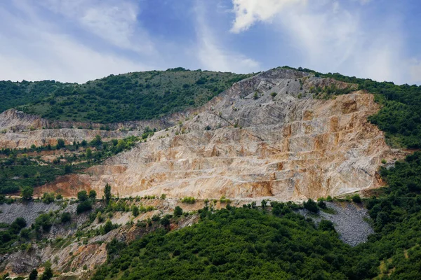 A natural stepped quarry rich in minerals is located near winding intermountain road, against backdrop of Rhodope Mountains and hills with extensive evergreen spruce forests and mountain vegetation