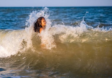 İki güzel bronzlaşmış uzun saçlı neşeli kız kardeş, parlak yaz mayo giyip, sıcak güneşli bir tatil akşamında, küçük deniz kabuklarıyla kumlu bir sürüde mavi köpüklü deniz dalgalarına atlarlar.