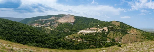 Panorama on industrial quarry for extraction of minerals with professional equipment and machines, and intermountain winding road passing by, in Rhodope Mountains covered with coniferous forests