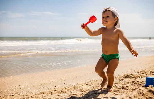 少し面白い子供 明るい休暇に暑い夏の太陽の下で砂の底に穏やかな青い海のシェルと小石を収集します — ストック写真