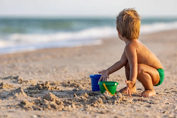 少し面白い子供 明るい休暇に暑い夏の太陽の下で砂の底に穏やかな青い海のシェルと小石を収集します — ストック写真