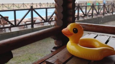 Sarı şişme bir ördek, dinlenme merkezindeki evlerden birinin konforlu çardağında, büyük bir havuz zeminine uzanan büyük, ahşap bir masanın üzerinde, sağanak yağmurla kaplı. 4K UHD yavaş çekim videosu