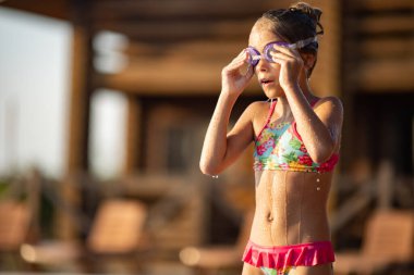 Parlak pembe mayo ve siyah yüzme gözlüğü takan bir kız, sıcak yaz güneşli günbatımının arka planında temiz şeffaf serin suyla derin mavi bir havuza atlıyor.