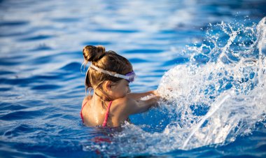 Parlak pembe mayo ve siyah yüzme gözlüğü takan bir kız, sıcak yaz güneşli günbatımının arka planında temiz şeffaf serin suyla derin mavi bir havuza atlıyor.