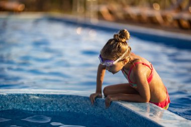Parlak pembe mayo ve siyah yüzme gözlüğü takan bir kız, sıcak yaz güneşli günbatımının arka planında temiz şeffaf serin suyla derin mavi bir havuza atlıyor.