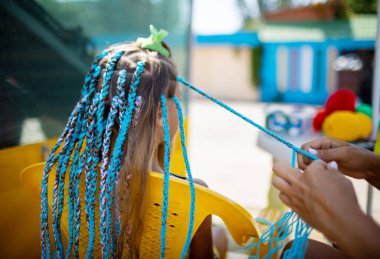 Parlak sarı bir yaz elbisesi içinde bronz tenli küçük bir kız, sıcak bir deniz gününde koyu ıslak saçlarıyla pembe-mavi Afrika örgüleri örüyor.
