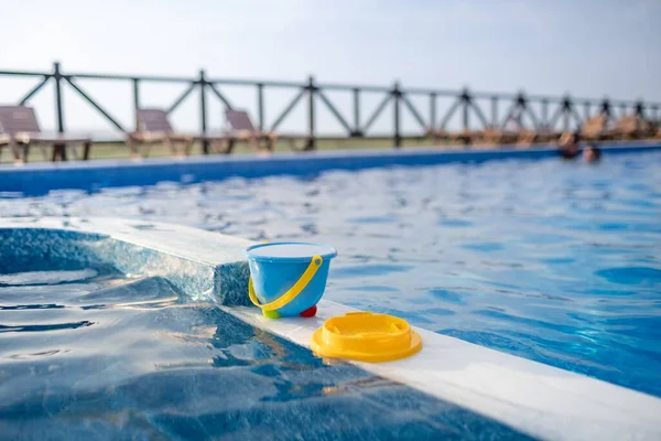 子供用の完全なバケツの水 黄色のふるい その他のビーチのおもちゃは 暖かい晴れた夏の日没を背景に透明な水で青いプールに乗っています — ストック写真