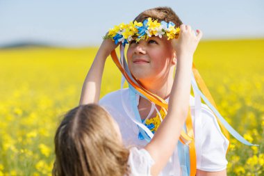 Nazik, genç kız kardeş, gülümseyen büyük erkek kardeşi, uzun renkli kurdeleli, parlak Ukrayna çelengini takıyor, sarı sınırsız tarlaların ve açık gökyüzünün arka planına karşı.