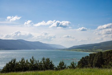 Işıl ışıl parlayan berrak bir göl, berrak mavi bulutlu bir gökyüzüne ve yuvarlanan yeşil ağaçlık dağ sıralarına ve küçük çiftçi dağ köylerine karşı parlak, ılık bir güneş ışığını yansıtır.