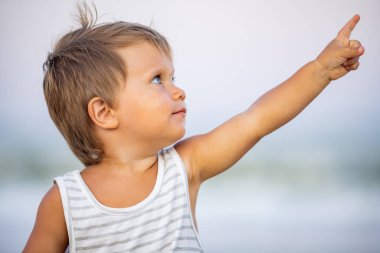 Parlak siyah-beyaz çizgili tişörtlü, açık sarı saçlı, sevimli, bronz tenli, kel bir çocuk. Büyük mavi gözlü, bulutların arasındaki uzaklığı gösteriyor. Dalgalı mavi denizin arka planında.