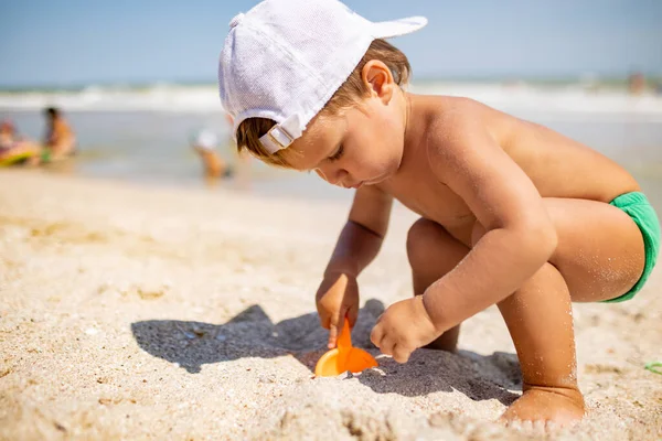 少し面白い子供 明るい休暇に暑い夏の太陽の下で砂の底に穏やかな青い海のシェルと小石を収集します — ストック写真