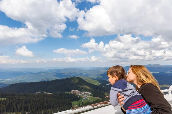Mother holds her baby son tightly and shows him the landscapes of mountain forest valley of the Rhodope Mountains and the blue cloudy sky, while standing on a high observation tower on top of Snezhan