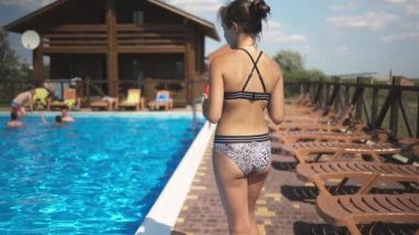 Siyah saçlı, parlak leopar mayosuyla topuz ören genç bir kız, yaz tatilinde güneşli bir günün tadını çıkaran çok renkli şişme bir topla havuz kenarında yürüyor.