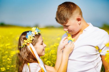 Beyaz elbiseli neşeli kız kardeş Ukrayna 'da çok renkli yaz kurdeleleri takıyor. Genç kardeşinin başında sarı tarlalar ve açık gökyüzü var.