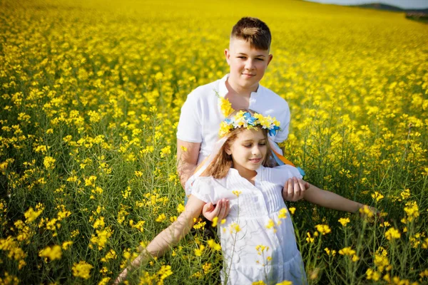 Big strong older teenage brother catches his little cheerful happy sister and bright Ukrainian flower wreath that falls into his hands, in rapeseed yellow flowering field under blue clear sky