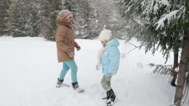 Side view of two pretty girls shaking pine tree in snowy forest. Happy caucasian sisters shaking snowy branch, smiling, enjoying snowfall with lush spruses on background. Concept of winter holidays.
