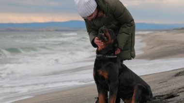 Sıcak ceketli kimliği belirsiz bir kız mavi fırtınalı denizin yanındaki kumlu sahilde duruyor ve sadık arkadaşının kulağının arkasını tırmalıyor. Rottweiler cinsinin büyük, eğitimli köpeği, 4K UHD yavaş çekim videosu.
