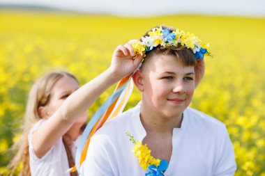 Beyaz elbiseli neşeli kız kardeş Ukrayna 'da çok renkli yaz kurdeleleri takıyor. Genç kardeşinin başında sarı tarlalar ve açık gökyüzü var.