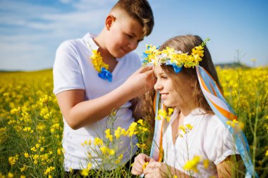 Önemseyen ağabeyi, parlak Ukrayna çiçek çelenkleriyle çok renkli ince kurdeleler örüyor. Küçük, gülümseyen kız kardeşin başında. Açık mavi gökyüzünün altında bitmeyen kolza tohumu tarlasında.