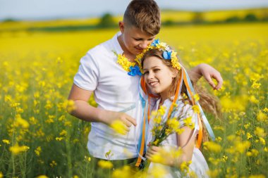 Mutlu kaygısız yerli çocukların ön görüntüsü: Genç erkek kardeş ve küçük kız kardeş, bulutlu gökyüzüne karşı sarı açan kolza tohumu tarlalarıyla çevrili yeşil çimlerle uzaklarda yürüyorlar.