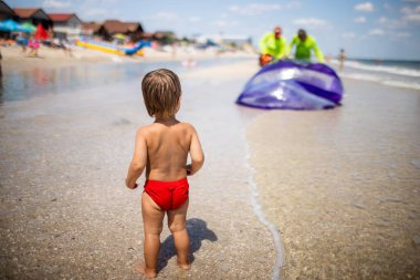 Sevimli, neşeli bir çocuk, mavi sakin deniz ufkuna bakıyor ve neşeli, neşeli bikinili çocuklar için parlak şişme toplar kavurucu güneşin altında kavurucu bir bikini giyiyor.