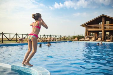 Parlak pembe mayo ve siyah yüzme gözlüğü takan bir kız, sıcak yaz güneşli günbatımının arka planında temiz şeffaf serin suyla derin mavi bir havuza atlıyor.