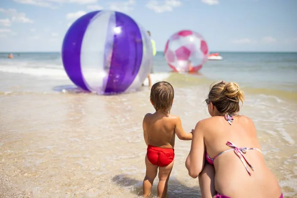 少しかわいい陽気な子供 青い穏やかな海の地平線と灼熱の太陽の下で明るいビキニで楽しい優しい幸せな母親と子供のための明るいインフレータブル大きなボールを見て — ストック写真