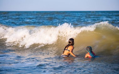 İki güzel bronzlaşmış uzun saçlı neşeli kız kardeş, parlak yaz mayo giyip, sıcak güneşli bir tatil akşamında, küçük deniz kabuklarıyla kumlu bir sürüde mavi köpüklü deniz dalgalarına atlarlar.