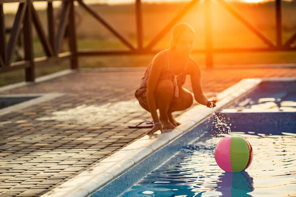 Fat tanned happy girl with dark hair braided in bun in bright summer suit is playing near pool with inflatable ball, against the background of golden summer evening sun