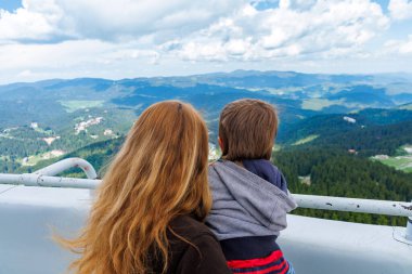 Anne, küçük oğlunu sıkıca tutuyor ve ona Rodop Dağları 'ndaki dağ ormanlarının manzaralarını ve Snezhan' ın tepesindeki yüksek bir gözlem kulesinde dururken mavi bulutlu gökyüzünü gösteriyor.