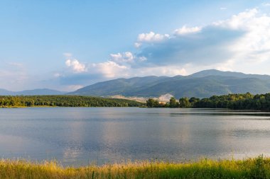Işıl ışıl parlayan berrak bir göl, berrak mavi bulutlu bir gökyüzüne ve yuvarlanan yeşil ağaçlık dağ sıralarına ve küçük çiftçi dağ köylerine karşı parlak, ılık bir güneş ışığını yansıtır.