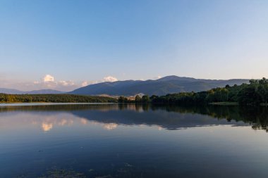 Işıl ışıl parlayan berrak bir göl, berrak mavi bulutlu bir gökyüzüne ve yuvarlanan yeşil ağaçlık dağ sıralarına ve küçük çiftçi dağ köylerine karşı parlak, ılık bir güneş ışığını yansıtır.