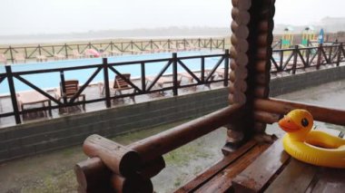 Sarı şişme bir ördek, dinlenme merkezindeki evlerden birinin konforlu çardağında, büyük bir havuz zeminine uzanan büyük, ahşap bir masanın üzerinde, sağanak yağmurla kaplı. 4K UHD yavaş çekim videosu