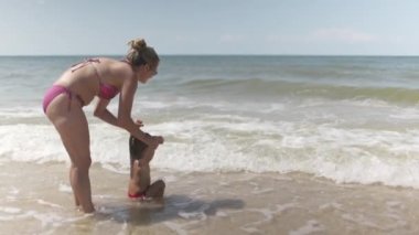 Neşeli, mutlu, şefkatli anne sıcak güneşin altında güneşli bir yaz tatilinde küçük komik oğlunu sakin mavi denizin üzerine fırlatıyor. 4K UHD yavaş çekim videosu