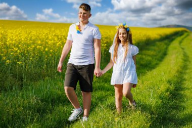 Mutlu kaygısız yerli çocukların ön görüntüsü: Genç erkek kardeş ve küçük kız kardeş, bulutlu gökyüzüne karşı sarı açan kolza tohumu tarlalarıyla çevrili yeşil çimlerle uzaklarda yürüyorlar.