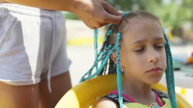 Parlak sarı bir yaz elbisesi içinde bronz tenli küçük bir kız, sıcak bir deniz gününde koyu ıslak saçlarıyla pembe-mavi Afrika örgüleri örüyor.