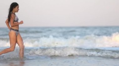 A fat dark-haired free girl with dark loose wet hair in a bright leopard swimsuit runs along the summer blue warm sea with foamy waves, peering into the distance of the sunny sea horizon