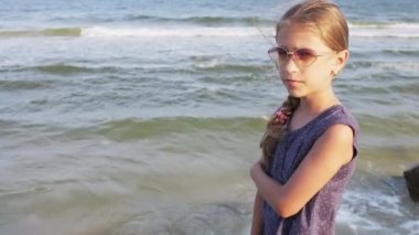A dreamy slim girl with blond hair braided in pigtail in light blue dress with small white polka dots and sunglasses looks thoughtfully into distance, standing on sea stone beach. HD slow-mo video