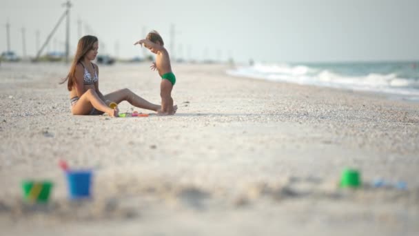 海岸近くの浅い海の水に座っている間 夏休みの暖かい晴れた日に 彼女の弟の幼児と一緒に遊んでいます Uhdスローモビデオ — ストック動画